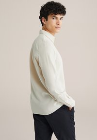 Dlhý rukáv, svetlo béžová košeľa s klasickým golierom a gombíkovými manžetami. Hladká textúra, mierne uvoľnený strih a zakrivený lem.