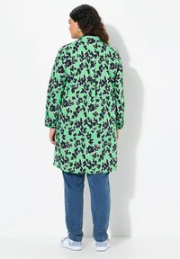 Tunika verde a maniche lunghe con stampa astratta nera, caratterizzata da una vestibilità ampia, orlo arrotondato e leggeri plissettature sul retro. Indossata con jeans blu.