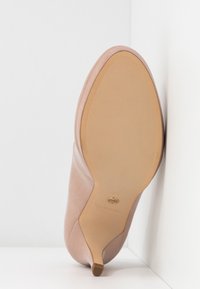 Chaussure à talon pour femme beige vue de la semelle, appuyée contre un mur d'angle blanc.