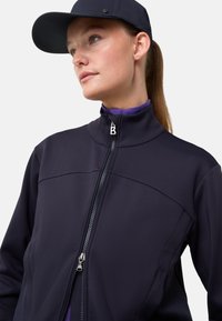 Námořnická softshellová bunda s vysokým límcem, celopropínacím zipem a kontrastním fialovým trikem pod ní. Čepice dodává sportovní nádech.