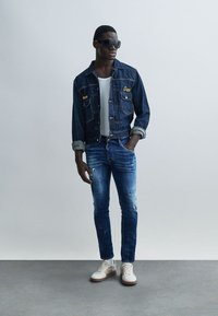 Spijkerjack met twee zakken, blauwe jeans met verweerde details, en witte sneakers met beige accenten. Zwarte zonnebril maakt de outfit compleet.