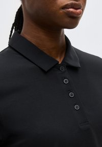 Κοντινό πλάνο ενός ατόμου που φοράει μαύρο πουκάμισο με γιακά και τέσσερα μαύρα κουμπιά, εμφανίζοντας τον λαιμό, το κάτω μέρος του προσώπου και την υφή του πουκάμισου.