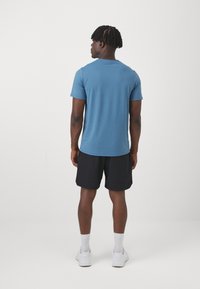 Μπλε κοντομάνικο μπλουζάκι συνδυασμένο με μαύρες φόρμες, απεικονιζόμενο από το πίσω μέρος. Το υλικό φαίνεται λείο, με casual σχέδιο. Φοράει λευκά αθλητικά παπούτσια.