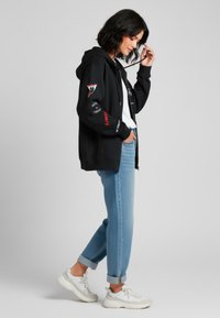 Felpa nera con zipper e dettagli grafici, t-shirt bianca sotto, jeans azzurri arrotolati e sneakers bianche. Texture liscia, vestibilità casual.
