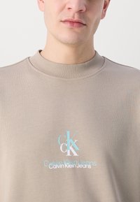 Béžová mikina s okrúhlym výstrihom vyrobená z bavlny, s rebrovaným golierom a viacfarebným vyšívaným logom v strede.