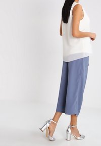 Top bianco senza maniche abbinato a pantaloni a gamba larga azzurri chiari e sandali con tacco quadrato argentati. I sandali hanno cinturini alla caviglia.