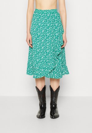 Jupe midi florale verte avec un ourlet froncé, ornée de motifs de fleurs blanches. Associée à des bottes de cowboy noires, ajoutant une texture contrastée.