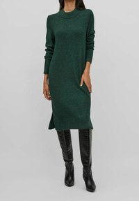 Abito verde lavorato a maglia con scollo rotondo, maniche lunghe, spacchi laterali e una vestibilità rilassata, abbinato a stivali neri al ginocchio con texture.