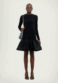 Čierne dlhé šaty s priliehavým telom a rozšírenou saténovou sukňou. Modelka drží malú károvanú kabelku a má na sebe čierne sandále na opätku.