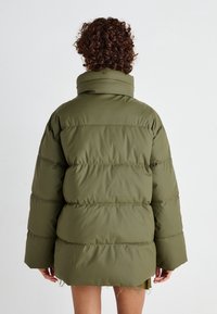 Chaqueta acolchada verde oliva con un cuello alto, que presenta mangas anchas y un diseño cuadrado. La parte posterior muestra paneles cosidos para aportar textura.