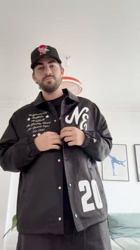 Svart jakke med hvit skrift og nummer "20." Har trykknapper, lange ermer og en glatt, blank tekstur.