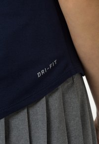 Marinblå Dri-FIT-tröja med fållen över en grå veckad kjol, med en del av en bar arm synlig på höger sida.