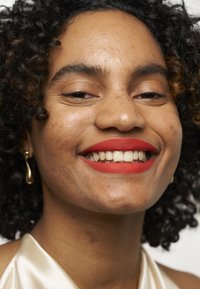 Ritratto di una persona con capelli neri ricci e lentiggini visibili, che indossa rossetto rosso e un top satin beige con un orecchino dorato.