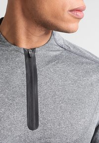 Top atlético gris con cierre de medio zip, textura de tela suave y mangas raglán con un sutil patrón jaspeado.