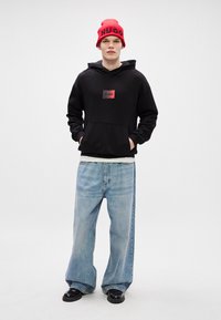 Μαύρη κουκούλα με αυτοκόλλητο λογότυπο, ανοιχτό μπλε χαλαρού στυλ τζιν παντελόνι και κόκκινο σκουφάκι με ριμπ που έχει κείμενο "HUGO". Μαύρα παπούτσια ολοκληρώνουν το σύνολο.