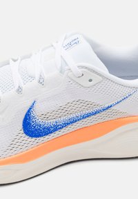 White athletic shoe with a textured mesh upper, blue gradient swoosh logo, and an orange accent on the sole. Features white laces and branding.