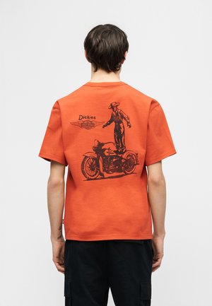 Άτομο που φοράει πορτοκαλί μπλουζάκι Dickies με μαύρη εικόνα ενός καουμπόη που ισορροπεί πάνω σε μια παλιά μοτοσικλέτα στην πλάτη.