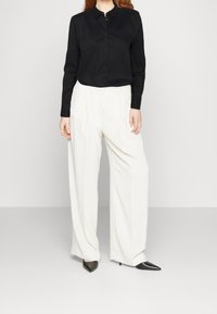 Camisa negra de manga larga con cuello abotonado, combinada con pantalones blancos de tiro alto y pernera ancha, y tacones negros de punta.