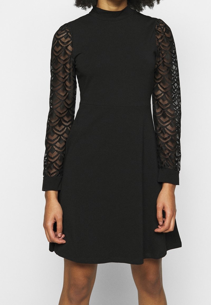 Femme portant une robe noire à col haut avec des manches longues transparentes à motifs et une jupe unie s'arrêtant au-dessus des genoux sur un fond uni.