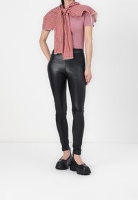 Person som bär svarta glänsande leggings, svarta kraftiga Mary Jane-skor, en mauve kortärmad topp och en röd rutig scarf över axlarna.