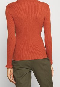 Czerwony sweter z prążkowanej tkaniny, z długim rękawem i dopasowanym krojem, zakończony falbaniastymi mankietami, połączony z oliwkowymi spodniami.