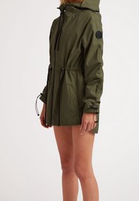 Femme portant une veste à capuche vert olive avec des cordons noirs et une fermeture éclair, montrée du milieu de la cuisse vers le haut sur un fond uni.