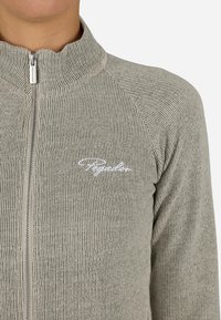 Kurtka ar rāvējslēdzēju, izgatavota no tonēta zaļa ribota auduma, ar augstu apkakli un baltu izšūtu logotipu uz krūtīm. Gluda tekstūra ar piegulošu dizainu.