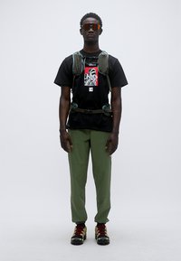 Μαύρο μπλουζάκι με γραφικό τύπωμα, πράσινο παντελόνι cargo και τσάντα πλάτης σε τυρκουάζ χρώμα. Κόκκινα και μαύρα αθλητικά παπούτσια με κίτρινες κορδόνια. Αντικατοπτριστικά γυαλιά ηλίου.