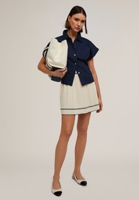 Camisa de botones de manga corta en azul marino, falda plisada color crema con rayas azul marino y zapatos de tacón bajo beige con punteras negras. Lleva un bolso blanco grande en la mano.