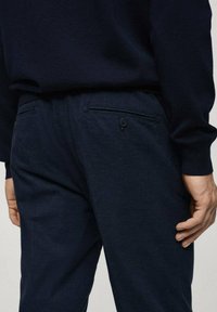 Pantaloni blu navy con una finitura testurizzata, dotati di una tasca posteriore con chiusura a bottone. La vita è elasticizzata per garantire comfort.