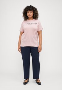 Svetloružová bavlnená tričko s čiernym textovým logom, v kombinácii s tmavomodrými nohavicami rovného strihu. Čierne ploché topánky dopĺňajú outfit.