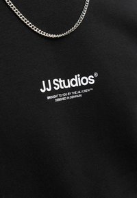 Camiseta de algodón negra con la marca blanca "JJ Studios" en el frente, acompañada de un collar de cadena plateado encima.