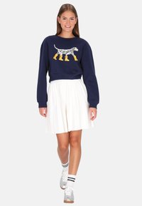 Tengerészkék pulóver, amelyen egy sárga esőcsizmát viselő dalmata figura látható, fehér pliszírozott szoknyával és fekete részletekkel díszített ezüst tornacipővel párosítva.