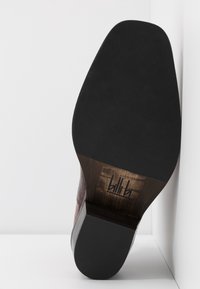 Semelle d'une chaussure habillée en cuir marron avec une semelle extérieure en caoutchouc noire et un talon en bois, appuyée en angle contre un mur blanc.