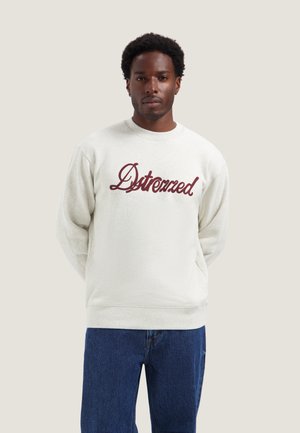 Man met kort krullend haar, die een wit sweatshirt draagt met "Distrezzed" in rode letters en een blauwe spijkerbroek, staand met handen achter de rug.