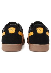 Zapatillas de ante negras con acentos amarillos, que presentan una suela de goma marrón texturizada y un logo dorado en el talón. Diseño minimalista.