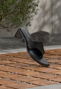 Μαύρο ψηλοτάκουνο σανδάλι Calvin Klein που αιωρείται πάνω από επιφάνεια με τούβλα και χαλίκια σε εξωτερικό χώρο, με θολό πράσινο φόντο.