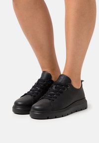Black leather sneakers feature a rounded toe, flat sole with small ridges, lace-up design, and minimal branding on the tongue.