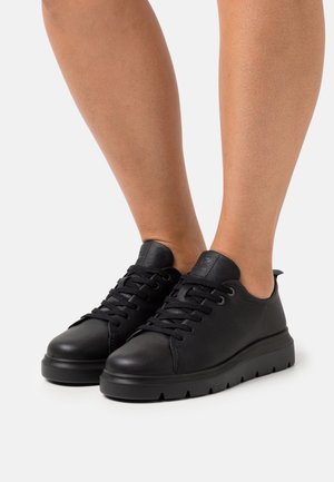 Las zapatillas de cuero negras presentan una puntera redondeada, suela plana con pequeñas estrías, diseño de cordones y un minimalista logo en la lengüeta.