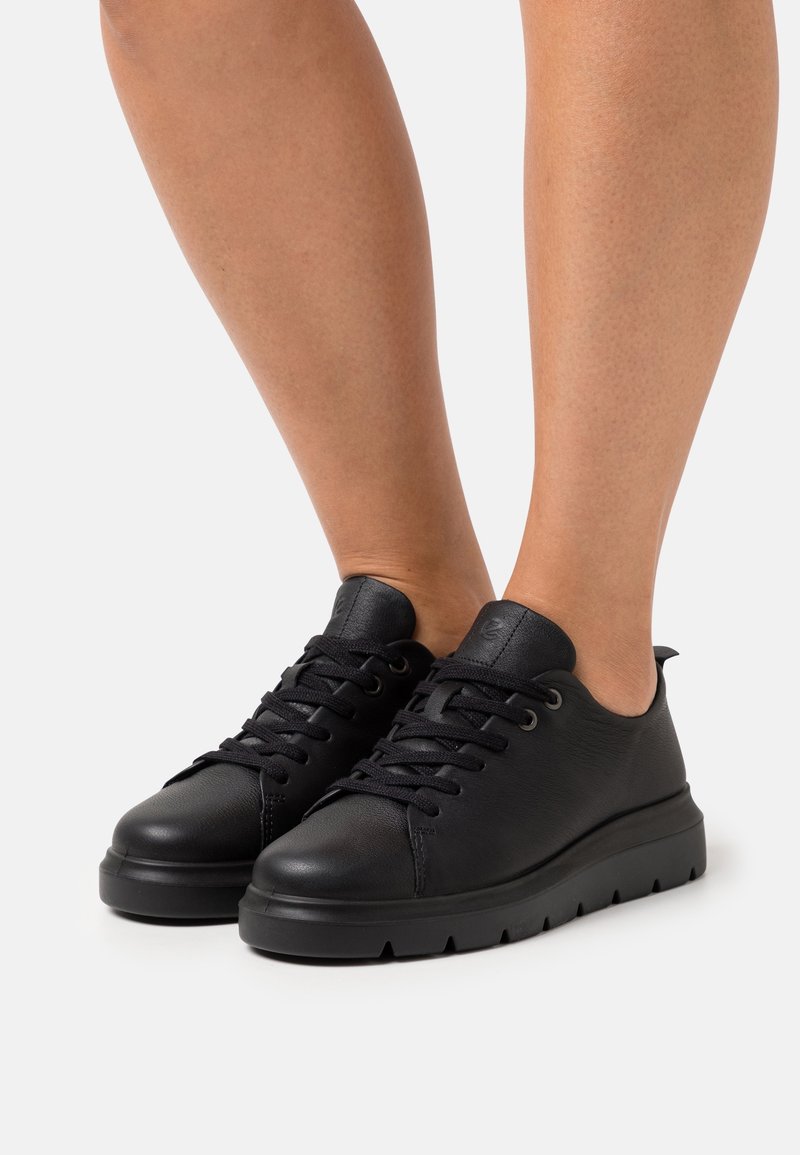 Black leather sneakers feature a rounded toe, flat sole with small ridges, lace-up design, and minimal branding on the tongue.