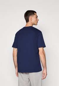 Mand iført en ensfarvet marineblå T-shirt med korte ærmer og lysegrå bukser, står med ansigtet mod en lysegrå baggrund og hovedet drejet mod højre.