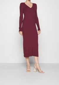 Burgundy ribbad klänning med lång ärm och V-ringning. Mittlång passform med en figurnära silhuett, tillsammans med beige blockklacksandaler.
