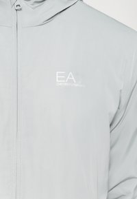 Šviesiai pilkas vėjui atsparus džemperis su gobtuvu, turintis užtrauktuką ir baltą "EA7 Emporio Armani" logotipą ant krūtinės. Lygi audinio tekstūra.