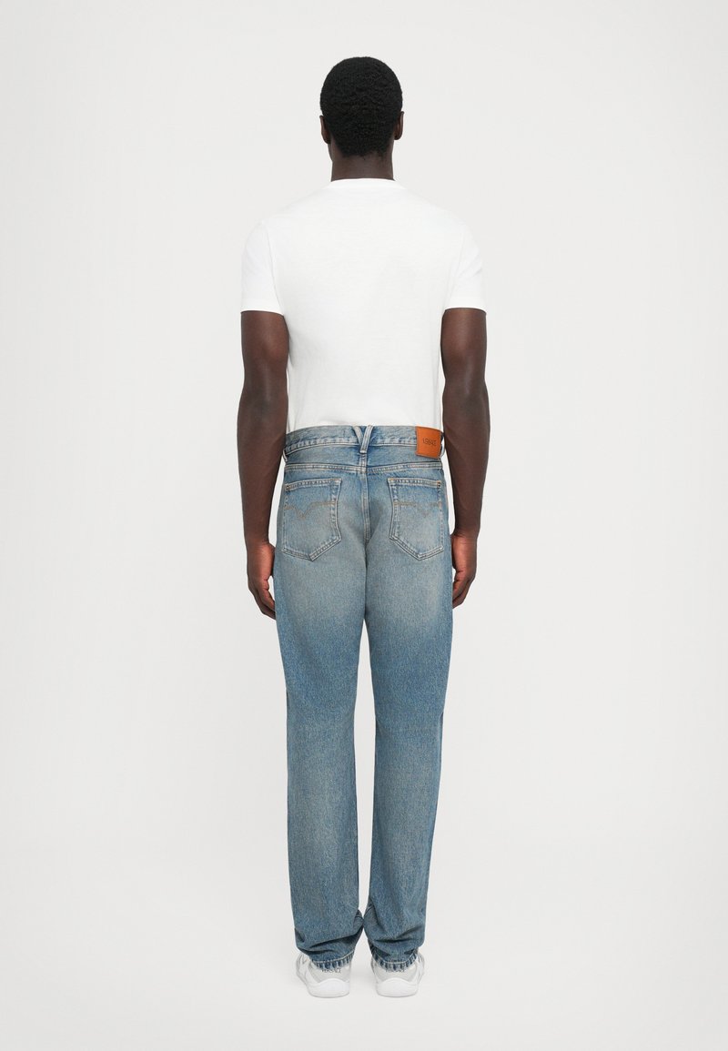 Man som står vänd bort, iklädd en vit t-shirt, ljusblå denimjeans med bakfickor, och vita sneakers mot en enfärgad bakgrund.