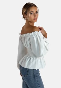 Weißes Off-Shoulder-Top mit Rüschenausschnitt, gerafftem Taillenbereich und fließenden Ärmeln. Kombiniert mit Jeans im Profilansicht.