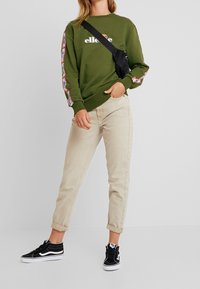 Sudadera verde oliva con logo en el centro, con detalles estampados en las mangas. Combinada con unos jeans beige claros con puños y zapatillas negras.
