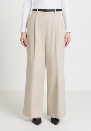 Pantalon beige à jambes larges avec plis à l'avant, une ceinture noire et une texture lisse. Stylisé avec des chaussures noires pointues.
