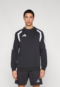 Μαύρο φούτερ με λευκές λεπτομέρειες, με στρογγυλή λαιμόκοψη, μακριά μανίκια και ριμπ μανσέτες. Το λογότυπο Adidas εμφανίζεται στο στήθος και στα σορτς.