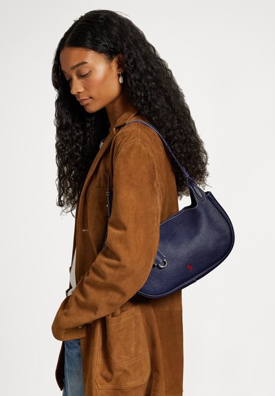 Ženska z dolgimi kodrastimi lasmi, oblečena v rjavo semiš usnjeno jakno in modre kavbojke, nosi temno modro teksturirano usnjeno torbo čez ramo z rdečim logotipom.