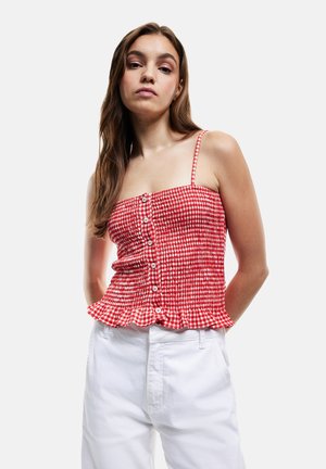Jeune femme aux longs cheveux portant un haut sans manches à carreaux rouges et blancs avec des boutons et un pantalon blanc sur un fond uni.